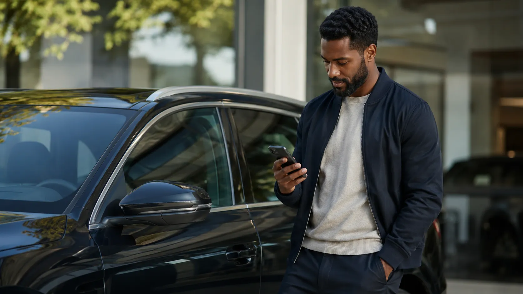 A buyer leans against their new car outside a dealership, checking their phone, quietly satisfied.
